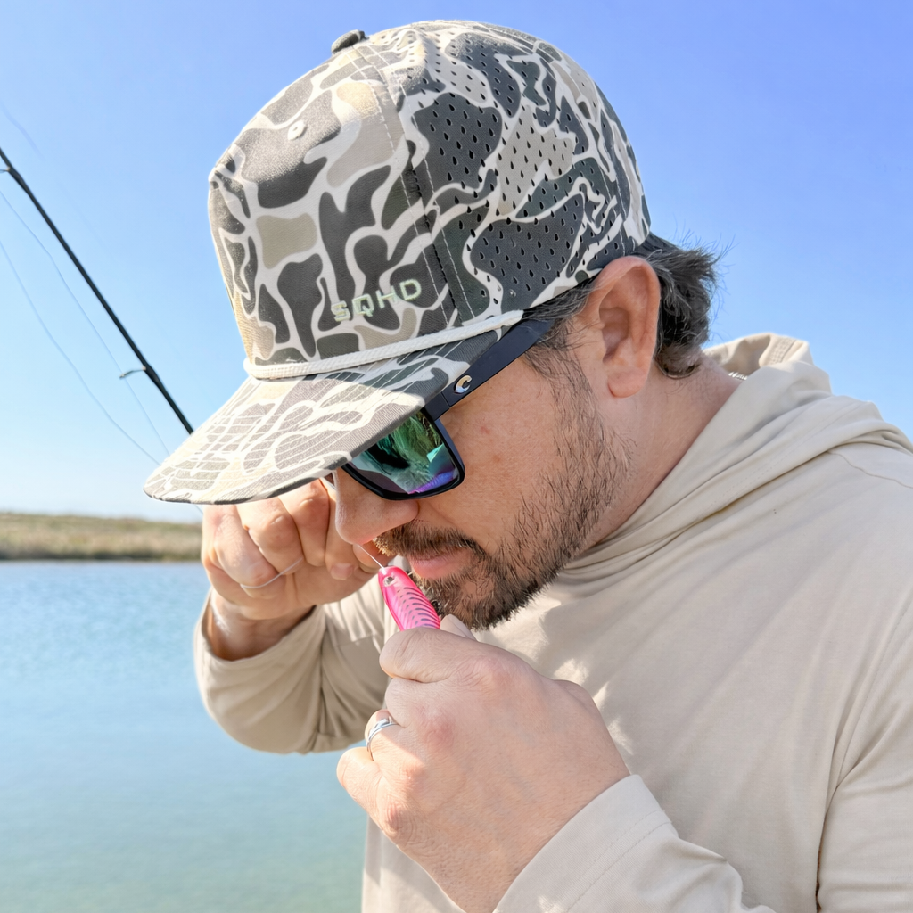 Man wearing a Salt SQHD Marsh Camo cap and sunglasses, tying on a pink lure by a body of water.